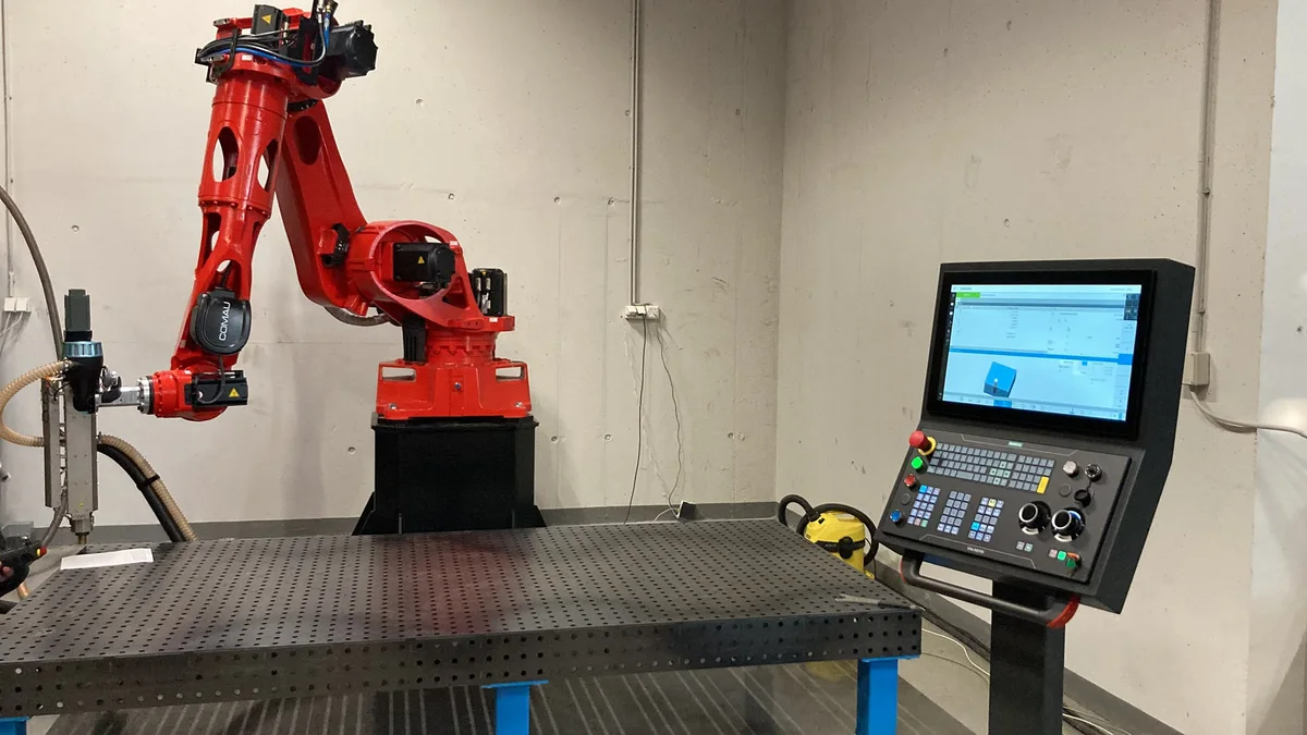 Červený priemyselný robot s tlačovou hlavou pri dierovanom oceľovom stole a riadiacom paneli.